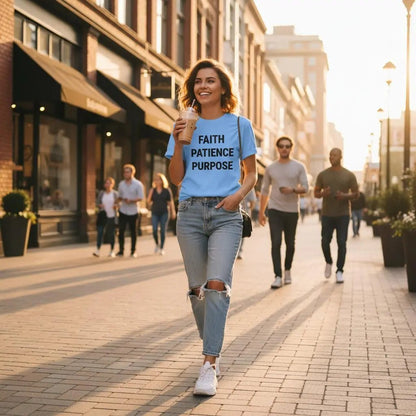 Woman wearing light blue t-shirt with FAITH PATIENCE PURPOSE text walking in sunlit city street